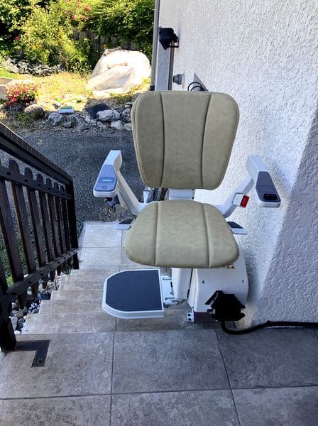 Monte‑escaliers extérieur avec siège rembourré beige monté sur un rail le long d’un escalier carrelé, accolé à la façade claire d’une maison avec jardin en arrière‑plan.