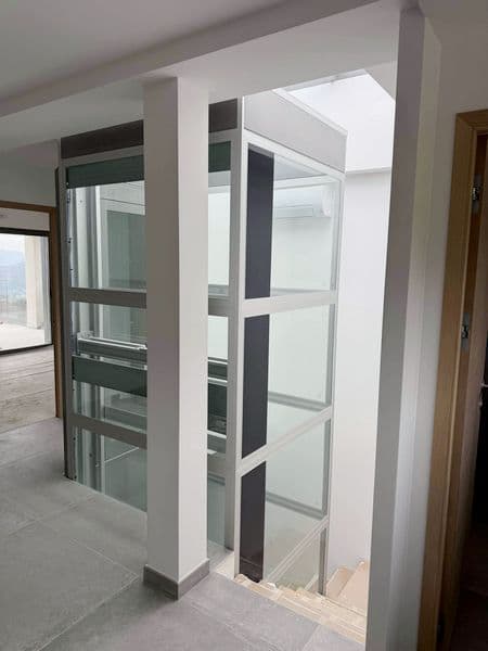 Ascenseur privatif vitré en structure grise, installé au centre d’un intérieur moderne avec cage transparente longeant un escalier en béton brut.