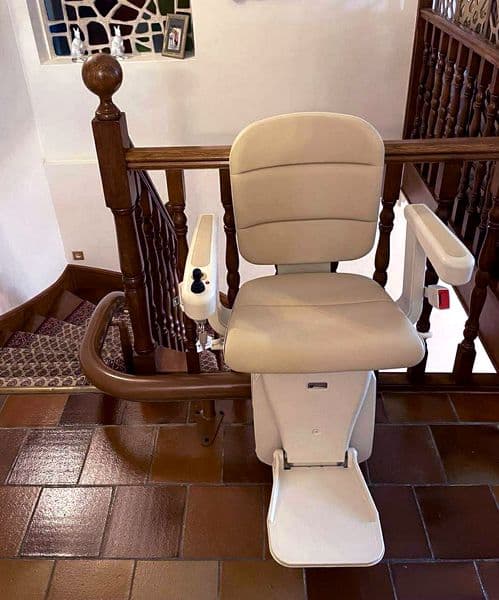 Monte‑escaliers intérieur avec siège rembourré beige en haut d’un escalier, installé sur un rail brun courbe fixé à la rampe en bois d’une maison.