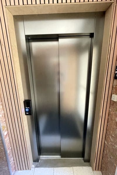 Cabine d’ascenseur moderne aux parois métalliques sombres, éclairage LED au plafond et panneau de commande tactile central, vue portes ouvertes depuis le palier.