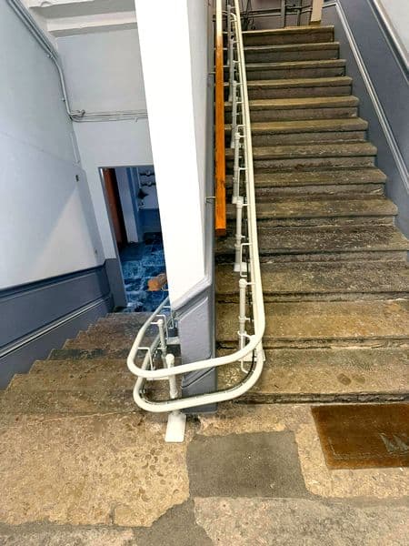 Monte‑escaliers courbe sans siège, montrant uniquement le rail tubulaire blanc qui longe un escalier ancien en pierre et contourne un angle de palier central dans un immeuble.